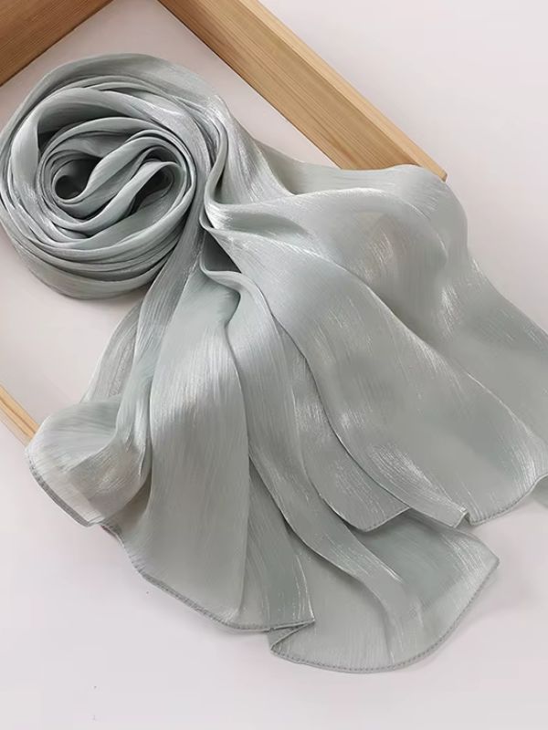 Hijab mousseline de soie froissée gris brume clair aux reflets shimmer argentés, présenté enroulé et déplié sur un plateau en bois naturel