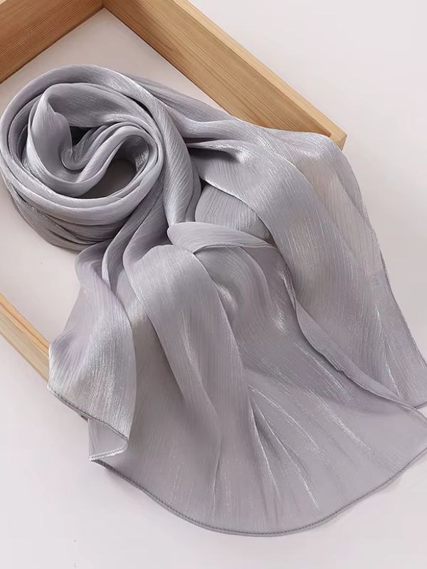 Hijab mousseline de soie froissée gris perle aux reflets shimmer argentés intenses, présenté enroulé et déplié sur un plateau en bois naturel