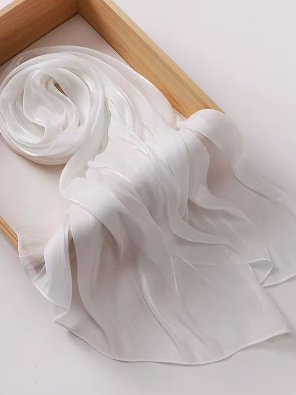 Hijab mousseline de soie froissée blanc nacré aux reflets lumineux, présenté déplié et enroulé sur un plateau en bois naturel