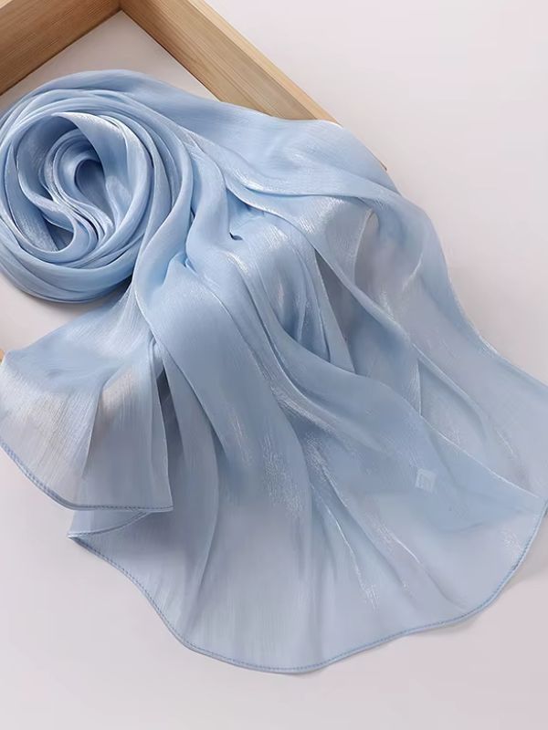 Hijab mousseline de soie froissée bleu ciel délicat aux reflets shimmer nacrés, présenté enroulé et déplié sur un plateau en bois naturel