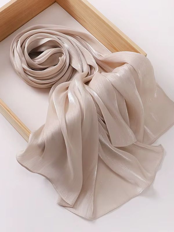 Hijab mousseline de soie froissée beige dune clair aux reflets shimmer nacrés, présenté enroulé et déplié sur un plateau en bois naturel