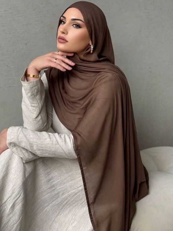 Femme portant un hijab jersey brun chocolat drapé élégamment, accessoirisée de bijoux dorés, assise dans une pose raffinée