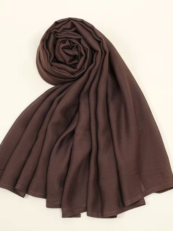 Femme portant un hijab jersey brun chocolat drapé élégamment, accessoirisée de bijoux dorés, assise dans une pose raffinée