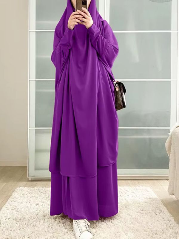 Une femme porte un jilbab violet ample et fluide dans un intérieur lumineux.