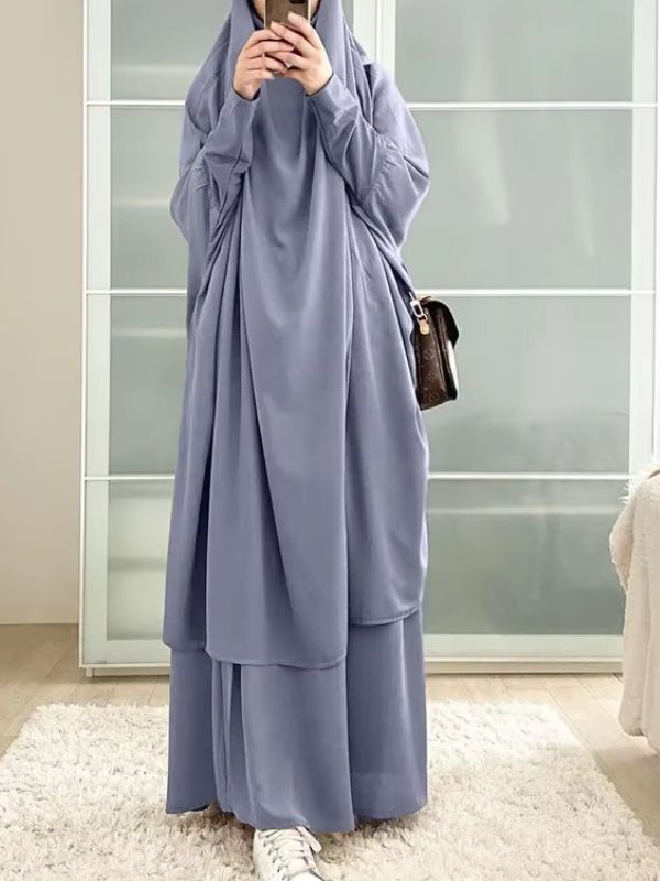 Une femme porte un jilbab gris clair ample et fluide dans un intérieur lumineux.