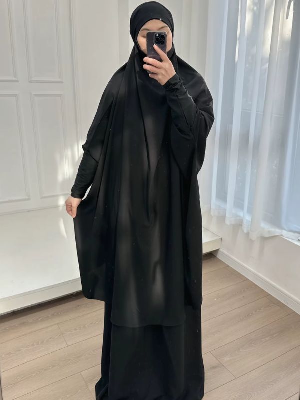 Jilbab femme deux pièces noir, grand haut à capuche et jupe longue, vue de face devant une fenêtre aux rideaux blancs