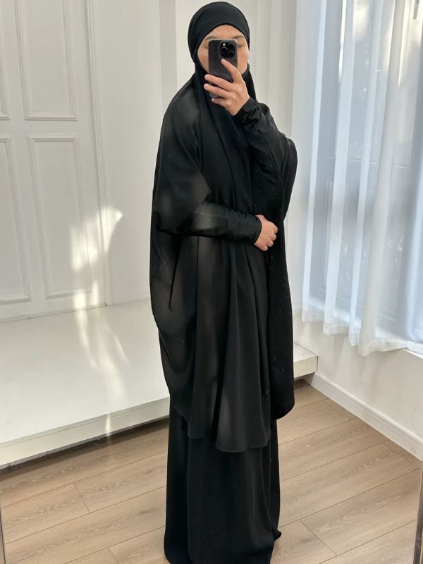 Jilbab femme noir vu de côté, grand haut ample à manches larges et jupe longue noire, main posée sur le haut