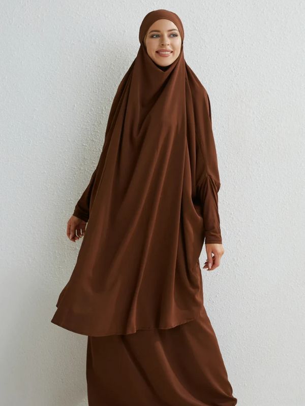 Femme souriante portant un jilbab long de couleur marron, posant devant un mur