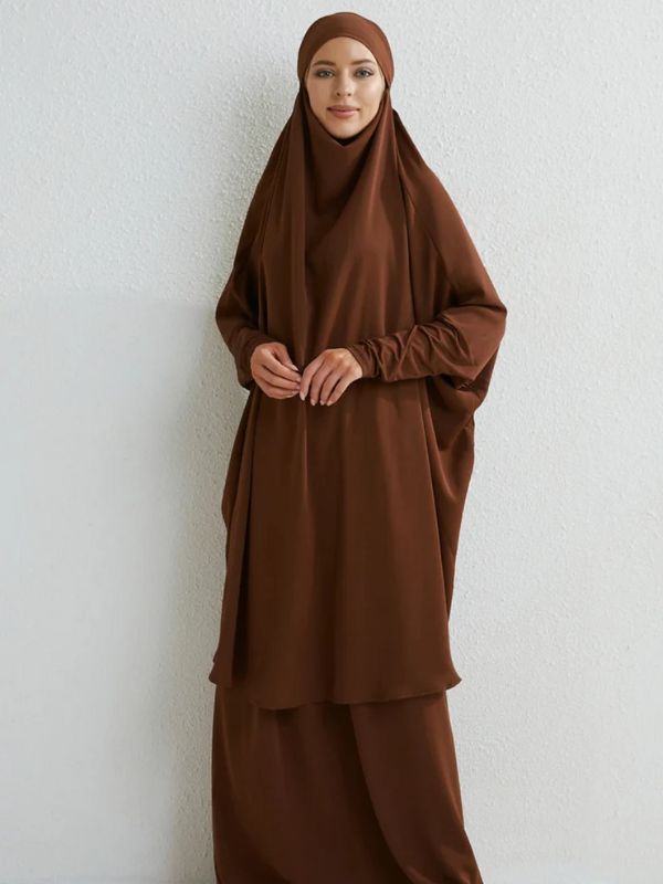 Femme souriante portant un jilbab long de couleur marron, posant devant un mur