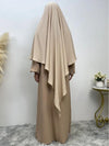 Femme portant un khimar long beige sable, vue de dos, posant dans un intérieur minimaliste avec des décorations naturelles et des fleurs en arrière-plan