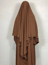 Femme en khimar marron devant un mur blanc et se tenant de dos