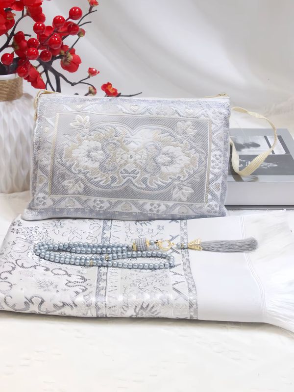 Tapis de prière Jasmin d'Argent plié, à motifs floraux blancs et argentés et franges, mis en scène avec un chapelet et une pochette