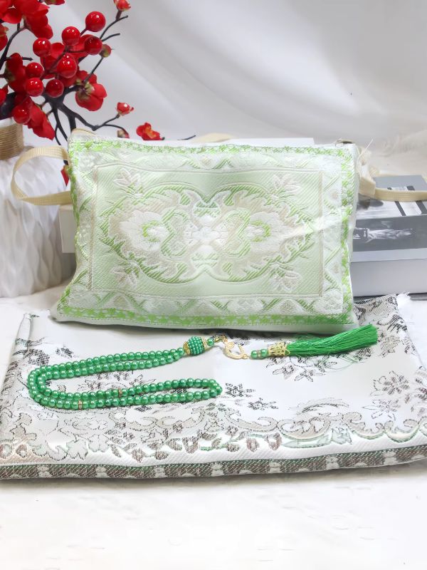 Tapis de prière Jasmin de Jade plié, fond blanc à motifs floraux gris taupe et touches de vert, mis en scène avec un chapelet vert et une pochette