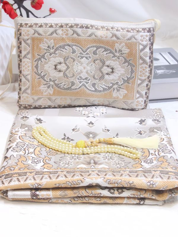 Tapis de prière Jasmin d'Ambre plié, fond gris perle à motifs floraux beige doré, sable et taupe, mis en scène avec un chapelet ivoire et une pochette