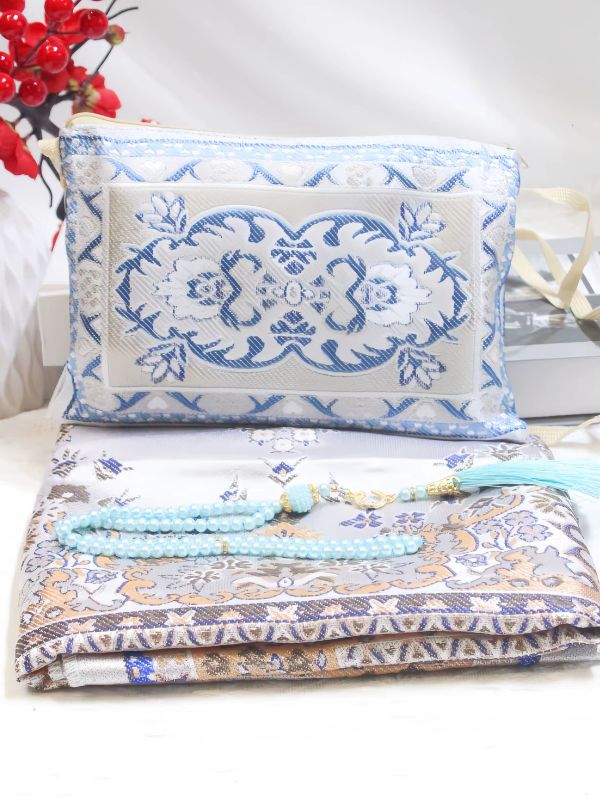 Tapis de prière Jasmin d'Azur plié, fond gris argenté à motifs floraux bleus et touches d'ocre, mis en scène avec un chapelet turquoise et une pochette